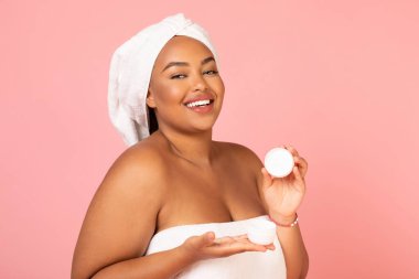 Happy Oversized African American Woman Showing Moisturizer Jar Advertising Skincare Product Standing Wrapped In Towel On Pink Background In Studio. Skin Care Cosmetics Concept