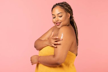 Bodycare. Attractive Overweight African American Lady Moisturizing Arm Caring For Skin Standing Wrapped In Towel Over Pink Studio Background. Skincare Cosmetics Concept