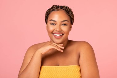 Portrait Of Pretty Obese Black Female Wrapped In Towel Smiling To Camera Posing Holding Hand Near Face Over Pink Studio Background. Beauty Routine And Wellness Concept