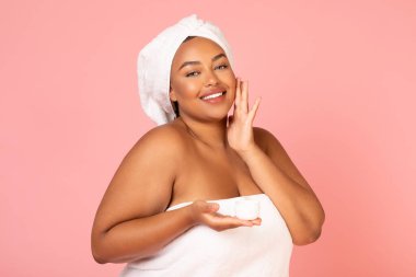 Plus Size Black Lady Holding Moisturizer Jar Applying Cream On Face Smiling To Camera Posing Wrapped In Towel On Pink Background, Studio Shot. Skincare Cosmetics Concept