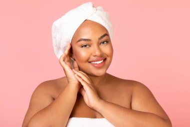 Happy Black Plus Size Female Holding Hands Near Face Caring For Skin Posing Wrapped In Towel Over Pink Studio Background, Smiling To Camera. Pampering And Beauty Care Concept