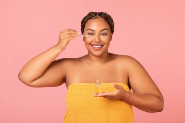 Cheerful Obese Black Woman Applying Facial Serum Moisturizing Face Skin Smiling To Camera Standing Posing Over Pink Studio Background. Beauty Routine And Spa Concept