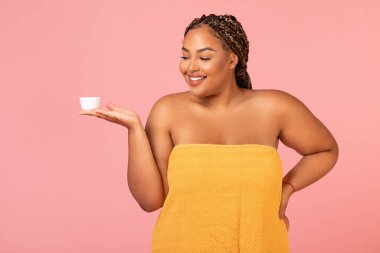 Pretty Obese Black Woman Holding Moisturizer Cream Jar Posing Wrapped In Towel Caring For Skin Standing Over Pink Studio Background. Facial Skincare Cosmetics, Beauty Routine Concept