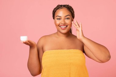 Facial Skin Care. Cheerful Overweight African American Lady Applying Moisturizer Holding Cream Jar On Face Smiling Posing On Pink Studio Background. Beauty Routine And Cosmetics Concept
