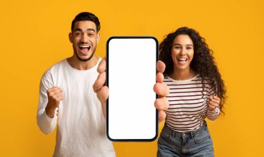 Happy emotional millennial arab couple enjoy online victory, show big smartphone with empty screen, isolated on yellow background, panorama, studio. Huge sale, new app, ad and offer, device for win