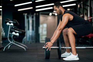 Young Black Male Athlete Adding Weight Plates On His Barbell, Motivated African American Sportsman Preparing For Weightlifting Workout, Exercising At Modern Sport Club Interior, Side View
