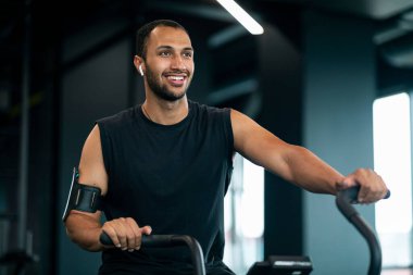 Young Black Male Athlete Training On Elliptical Bike Machine At Gym, Portrait Of Smiling African American Sportsman Exercising In Modern Fitness Club Interior, Enjoying Healthy Lifestyle, Copy Space