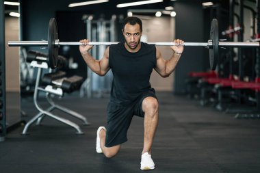 Athletic Young African American Man Making Squats With Heavy Barbell At Gym, Motivated Black Sportsman Go Down On One Knee And Looking At Camera, Making Bodybuilding Training At Modern Sport Club