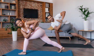 Smiling young african american male and female in sportswear doing exercises for legs or practicing yoga in living room interior, panorama. Body care together, weight loss, fit at home during covid-19