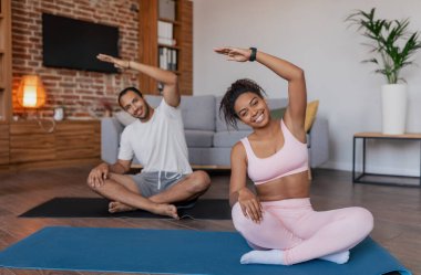 Cheerful young african american male and woman in sportswear do stretching, workout and practice yoga on mat in living room interior. Weight loss, body care and fitness at home due covid-19 quarantine
