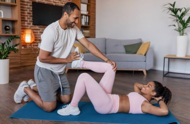 Glad young african american male help to female in sportswear do leg exercises on mat in living room interior. Stretching, support for body care, fitness and workout at home, new normal due covid-19