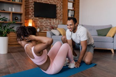 Glad young african american man help to lady in sportswear doing abs exercises on mat, enjoy workout in living room interior. Support with weight loss together, fitness and exercises at home, covid-19