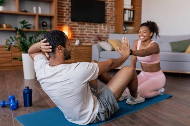 Cheerful young african american husband and wife in sportswear doing abs exercises on mat together in living room interior. Weight loss, fitness and workout at home, body care due covid-19 quarantine