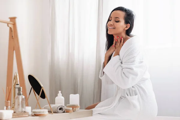 Pretty Mixed Woman Touching Neck Caring For Skin Looking At Mirror Enjoying Beauty Routine Sitting In Modern Bathroom Indoors. Skincare Cosmetic Products Concept