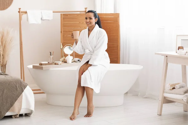 Morning Beauty Routine. Happy Mixed Woman Drinking Coffee Enjoying Selfcare Smiling To Camera Sitting On Bathtub In Modern Bathroom Indoors, Wearing Bathrobe. Spa And Wellness Concept