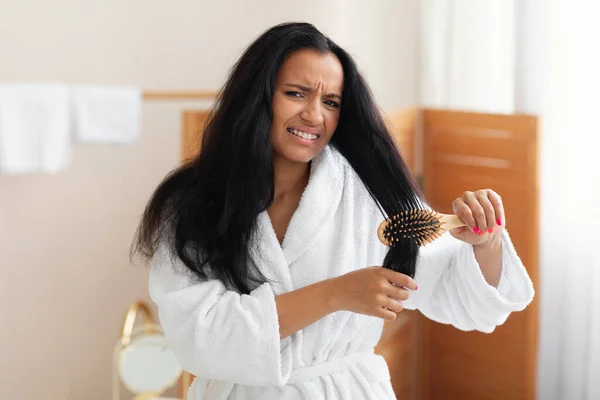 Unhappy Woman Having Problem Brushing And Detangling Tangled Long Hair Using Wooden Hairbrush In Modern Bathroom At Home, Looking At Camera. Haircare Treatment Concept