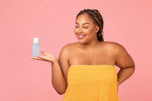 Skincare Cosmetics. Oversized African American Female Holding Micellar Water Bottle Advertising Cosmetic Product Standing Posing Wrapped In Towel Over Pink Background. Studio Shot