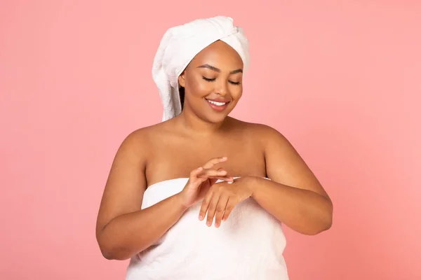 Skin Care Cosmetics. Happy Oversized African American Female Applying Moisturizer Cream On Hands Standing Wrapped In Towel On Pink Studio Background. Beauty And Pampering Concept
