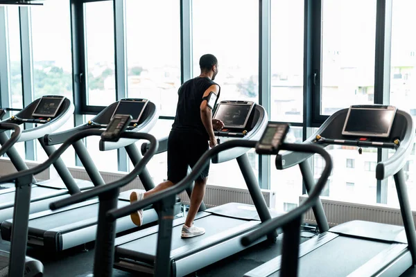 Cardio Training. Young Black Man Running On Treadmill Near Panoramic Window At Gym, Back View Of Sporty African American Male Jogging With Modern Equipment At Fitness Club, Copy Space