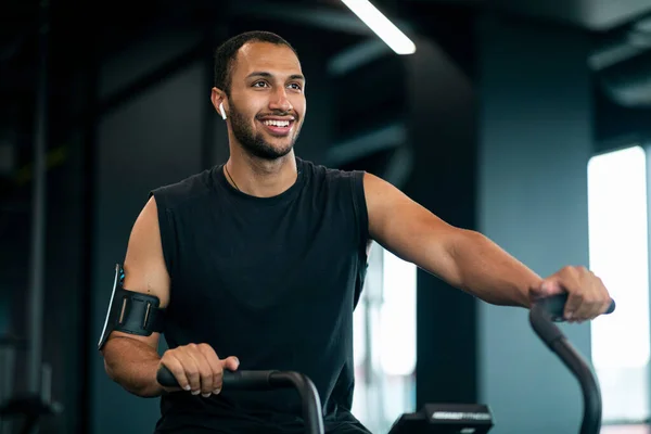 Young Black Male Athlete Training On Elliptical Bike Machine At Gym, Portrait Of Smiling African American Sportsman Exercising In Modern Fitness Club Interior, Enjoying Healthy Lifestyle, Copy Space