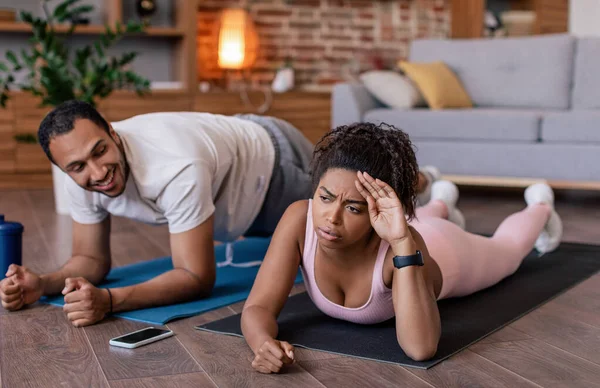 Smiling young black man supports tired sad wife in sportswear do plank exercises on mat with bottle of water and phone in room interior. Overwork, weight loss together, problems with workout at home