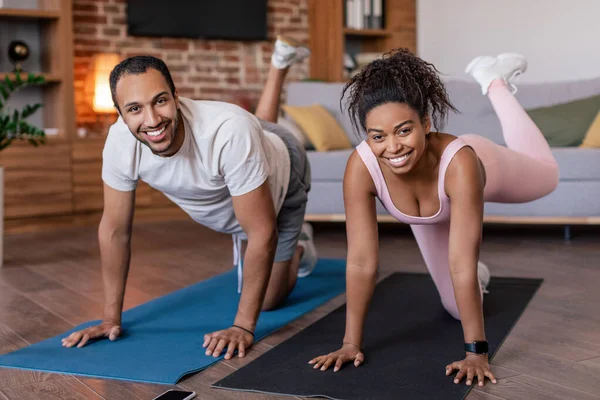 Glad young african american male and female in sportswear doing leg exercises on mat in living room interior. Workout for weight loss, body care together, fitness at home during covid-19 quarantine