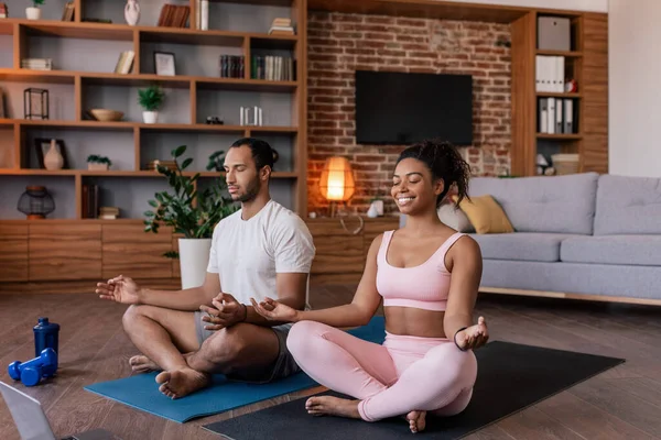 Happy calm young african american husband and wife in sportswear meditate or practice yoga on mat with dumbbells in living room interior. Body care together, fitness and workout at home due covid-19