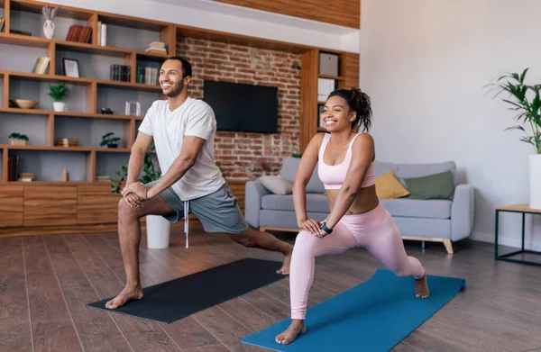 Glad young african american couple athletes in sportswear do exercises or practicing yoga in living room interior. Fitness for muscle, meditation at home, weight loss, body care together due covid-19