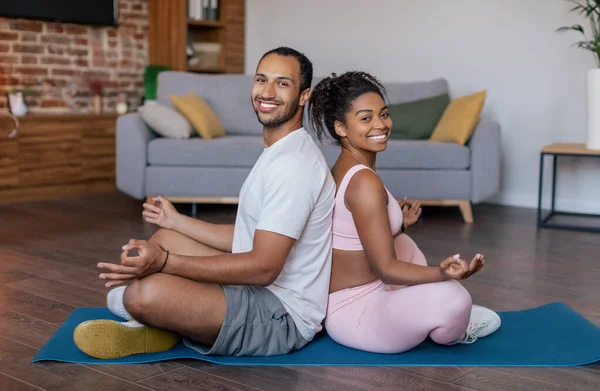 Happy young african american guy and lady in sportswear practice yoga, meditation, back to back on mat in living room interior. Body care, weight loss and workout at home gym due covid-19 quarantine