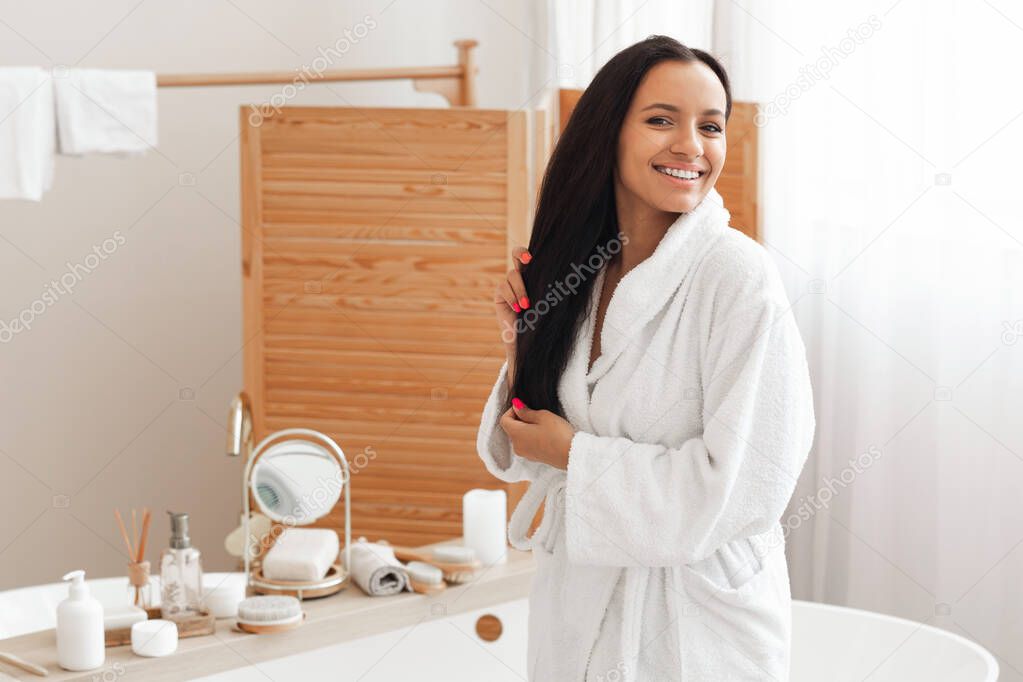 Haircare. Cheerful Mixed Lady Posing Touching Long Brunette Hair Smiling To Camera In Modern Bathroom Indoors, Wearing Bathrobe. Beauty Routine And Cosmetics Concept