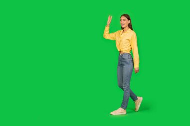 Cheerful Lady Walking Waving Hand Looking Aside At Free Space Over Green Studio Background. Full Length Shot Of Woman Gesturing Hello. Hi, Nice To Meet You Concept. Side View