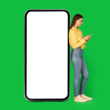 Cheerful Woman Texting On Smartphone Leaning On Large Phone With Empty Screen In Studio Over Green Background. Great Mobile Application Advertisement. Mockup, Square