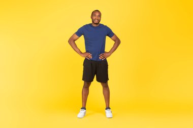 Sporty Black Male Posing Smiling To Camera Holding Hands On Hips Standing In Studio On Yellow Background, Wearing Sportswear. Workout Training Concept. Full Length