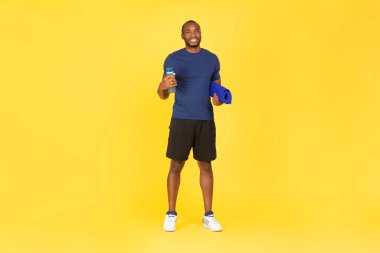 Sporty African American Male Holding Water Bottle And Fitness Mat Posing Ready For Training Standing Over Yellow Background. Workout Equipment Concept. Studio Shot, Full Length