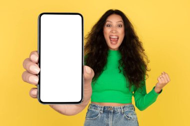 Emotional Arabic Lady Showing Huge Phone With Blank Screen Shouting And Gesturing Yes Posing Standing Over Yellow Background In Studio. Victory, Great Mobile News Concept. Mockup
