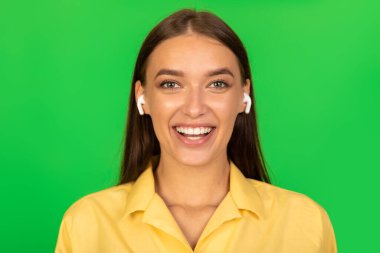 Headshot Of Joyful Lady Wearing Earbuds Earphones Listening To Music Smiling To Camera Posing Over Green Studio Background. Great Musical Application And Playlist Concept