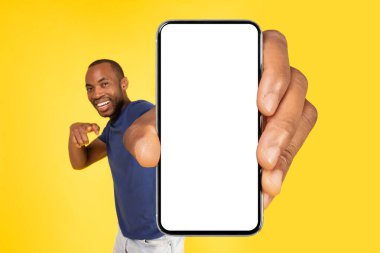 Wow App. Cheerful Black Guy Showing Cellphone With Huge Empty Screen And Pointing Finger Recommending Great Application Over Yellow Studio Background. Mobile Offer Advertisement. Mockup