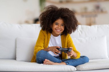 Happy preteen african american kid girl with bushy hair sitting on couch, holding joystick and smiling, carefree child playing video game at home, copy space. Kids and modern technologies concept