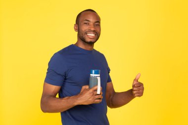 Fit Black Man Holding Fitness Bottle Gesturing Thumbs Up Approving Workout Smiling To Camera Standing Over Yellow Studio Background. Sport And Healthy Hydration Concept
