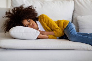 Upset little black girl with curly hair in casual outfit lying on pillow on couch alone, spending time at home, feeling lonely and bored, longing her friends and parents, copy space