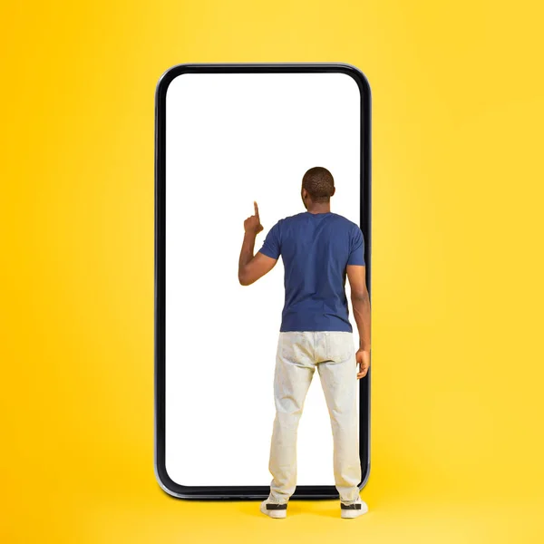 Black Man Using Huge Phone With Blank Screen Standing Back To Camera On Yellow Studio Background. Male Using Great Application Advertising Mobile Offer Concept. Full Length, Square