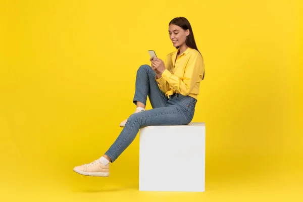 Happy Female Using Smartphone Websurfing And Texting Sitting On Cube In Studio Over Yellow Background. Lady Using Mobile Application And Networking In Social Media Via Cellphone. Side View