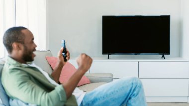 Overjoyed black man watching sports game on TV, celebrating victory and shaking fists, sitting on sofa, looking at empty screen, mockup, selective focus