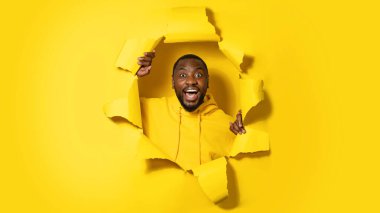 Excited african american man peeking out of hole in torn yellow wall background, wearing hoodie and looking at camera with open mouth, banner, copy space