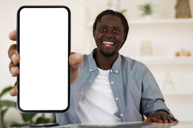 Online Ad. Cheerful Black Guy Demonstrating Smartphone With Big Blank Screen While Sitting At Desk At Home, African American Man Recommending New Mobile App Or Website, Collage, Mockup
