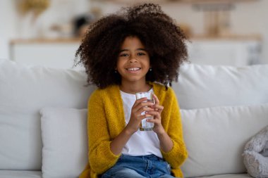 Cheerful pretty little black girl with bushy hair sitting on couch at home, holding glass of water and smiling at camera, kid drinking water, copy space. Hydration, children healthy lifestyle