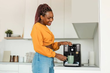 Excited young black woman making fresh aromatic coffee in modern machine in kitchen interior, enjoying hot morning beverage at home, free space