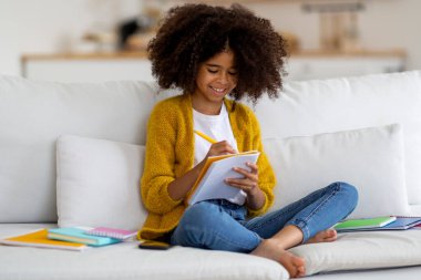 Happy african american school girl with bushy hair sitting on couch, holding notepad and pen, writing essay and smiling, kid doing homework, enjoying educational process, home interior, copy space