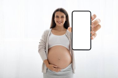 Cheerful young long-haired pregnant woman in homewear showing cell phone with white empty screen and smiling, recommending nice mobile app, white background, mockup for advertisement, copy space