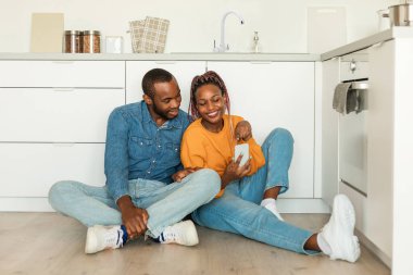 Positive african american man and woman looking at smartphone, sitting on floor at kitchen interior, copy space. Great offer, new blog, device and app, advertisement and sale